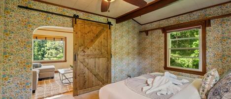 A bedroom with a bed, a ceiling fan, a wooden door, a window, and a view of greenery outside.