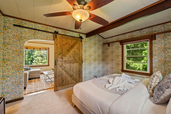 A bedroom with a bed, a ceiling fan, a wooden door, a window, and a view of greenery outside. - Storybook Upper Suite | Pool, Hot Tub, BBQ Grill (Seattle)