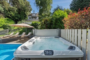 A hot tub with a control panel, surrounded by a pool, lounge chairs, and a wooden fence.