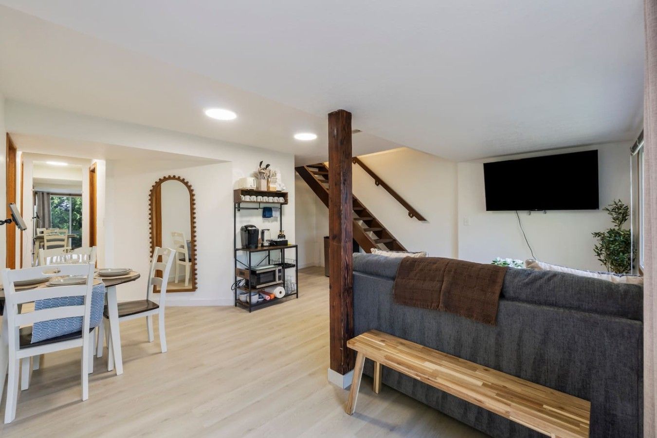 A living room with a grey sofa, wooden bench, dining table with chairs, a mirror, and a shelf with various items.