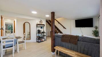 A living room with a grey sofa, wooden bench, dining table with chairs, a mirror, and a shelf with various items.