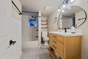 A modern bathroom with a bathtub, toilet, vanity with drawers, and a large mirror with lights. - Cozy 1BR/1BA Lower Unit Near UW | Walk to Campus (Seattle)
