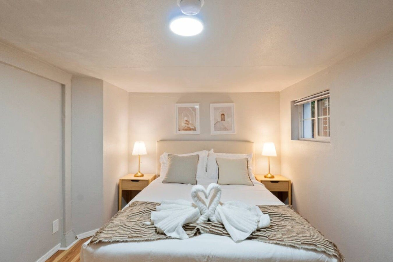 A hotel room with a bed, two bedside lamps, a window, and two framed pictures on the wall.