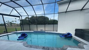 Una piscina con cerramiento de vidrio, rodeada por un solárium empedrado y un edificio blanco.