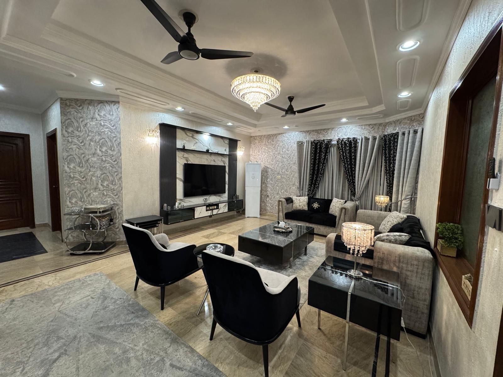 A modern living room with a flat-screen TV, black and grey sofas, a coffee table, and a chandelier.