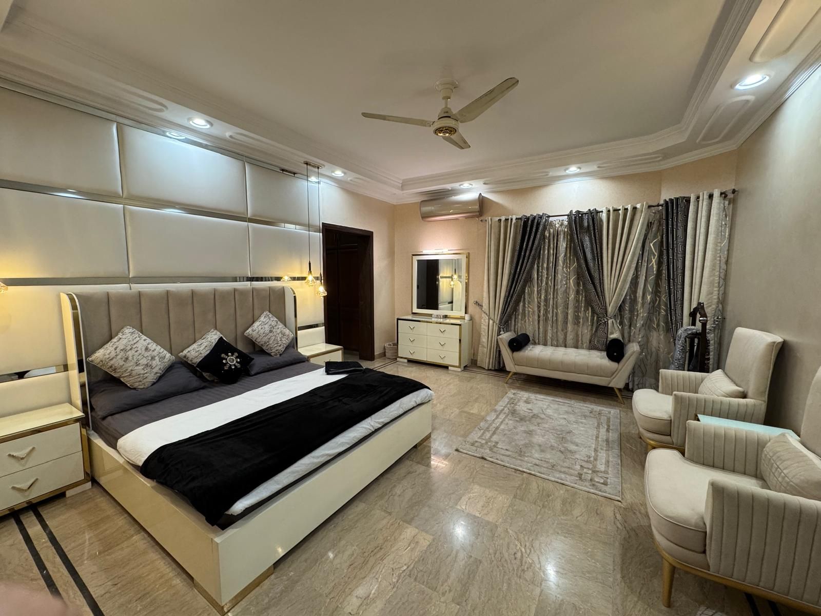 Une chambre moderne avec un grand lit, des tables de chevet, une coiffeuse, un ventilateur de plafond et un coin salon agrémenté de rideaux.