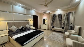 Une chambre moderne avec un grand lit, des tables de chevet, une coiffeuse, un ventilateur de plafond et un coin salon agrémenté de rideaux.