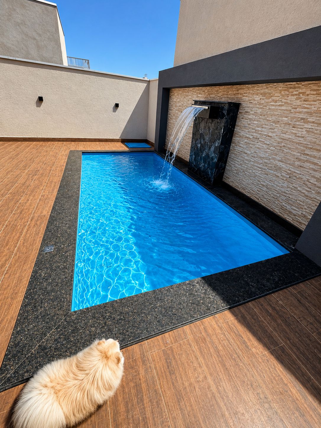 A rooftop swimming pool with a waterfall feature, surrounded by wooden decking and stone walls.