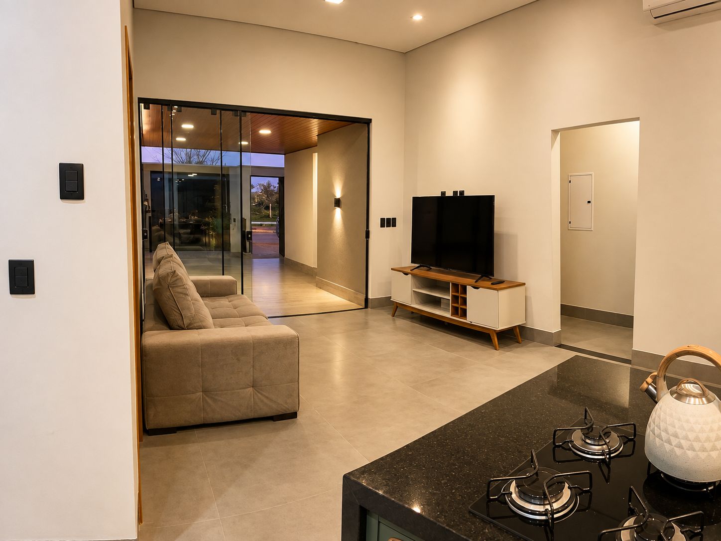 A modern living room with a sofa, a flat-screen TV on a wooden stand, and a kitchen area with a gas stove.