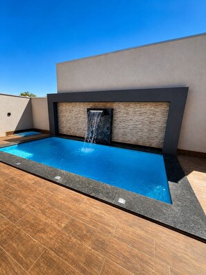 A modern outdoor swimming pool with a waterfall feature, surrounded by a wooden deck and a stone wall. - BONITO CASA MIRRA - Cozy house in Bonito with pool and space for family. (Bonito)