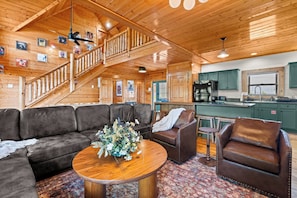 Ein gemütliches Blockhaus-Interieur mit einer Holztreppe, einem Wohnbereich mit einer Couch und Sesseln, einer Küche mit grünen Schränken und einem Deckenventilator.