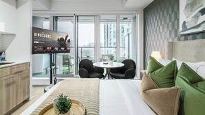 Ein modernes Hotelzimmer mit einem großen Bett, einem Essbereich, einem Fernsehbildschirm, der 'The Dinosaurs' zeigt, und Blick auf die Stadt durch große Fenster.