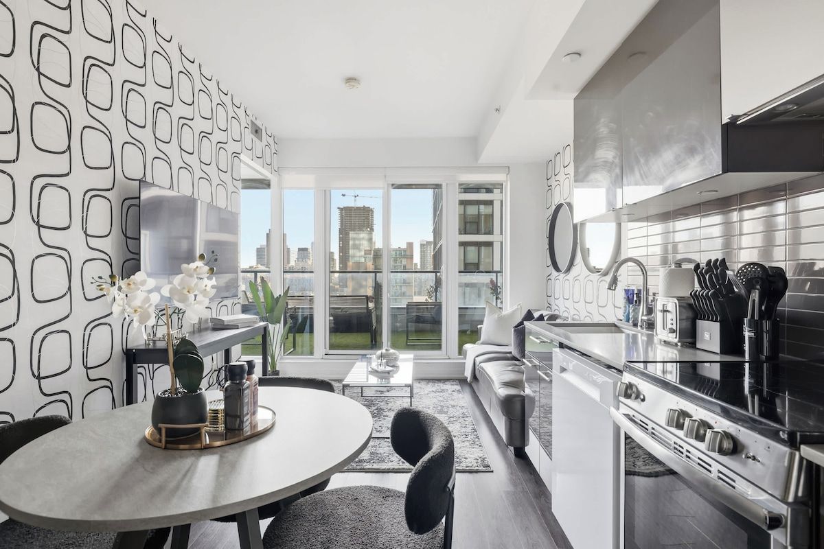 A modern kitchen with a built-in oven, a dining area with a round table, and a large window offering a city view.