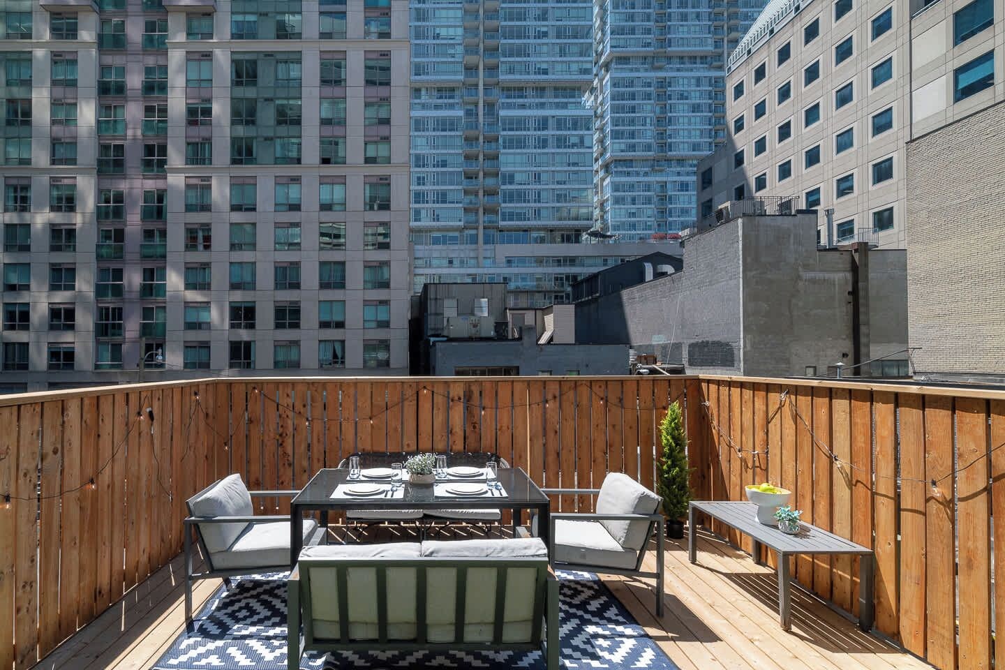 A rooftop patio with a dining table set for four, surrounded by wooden railings and a patterned rug.