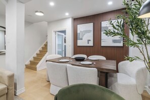 Un coin repas avec une table en bois, des chaises blanches et un escalier en arrière-plan.