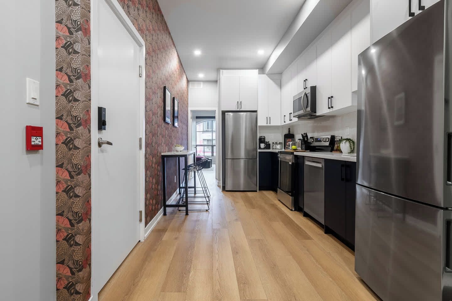 A modern kitchen with stainless steel appliances, a wooden floor, and a patterned wallpaper on the left wall.