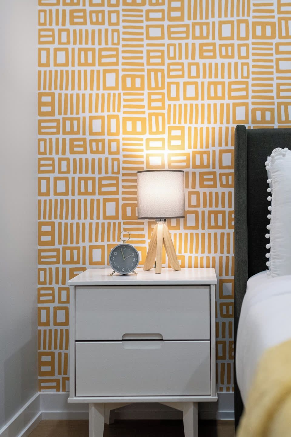 Une table de chevet avec une lampe et un réveil, un tiroir, et un lit avec une tête de lit.
