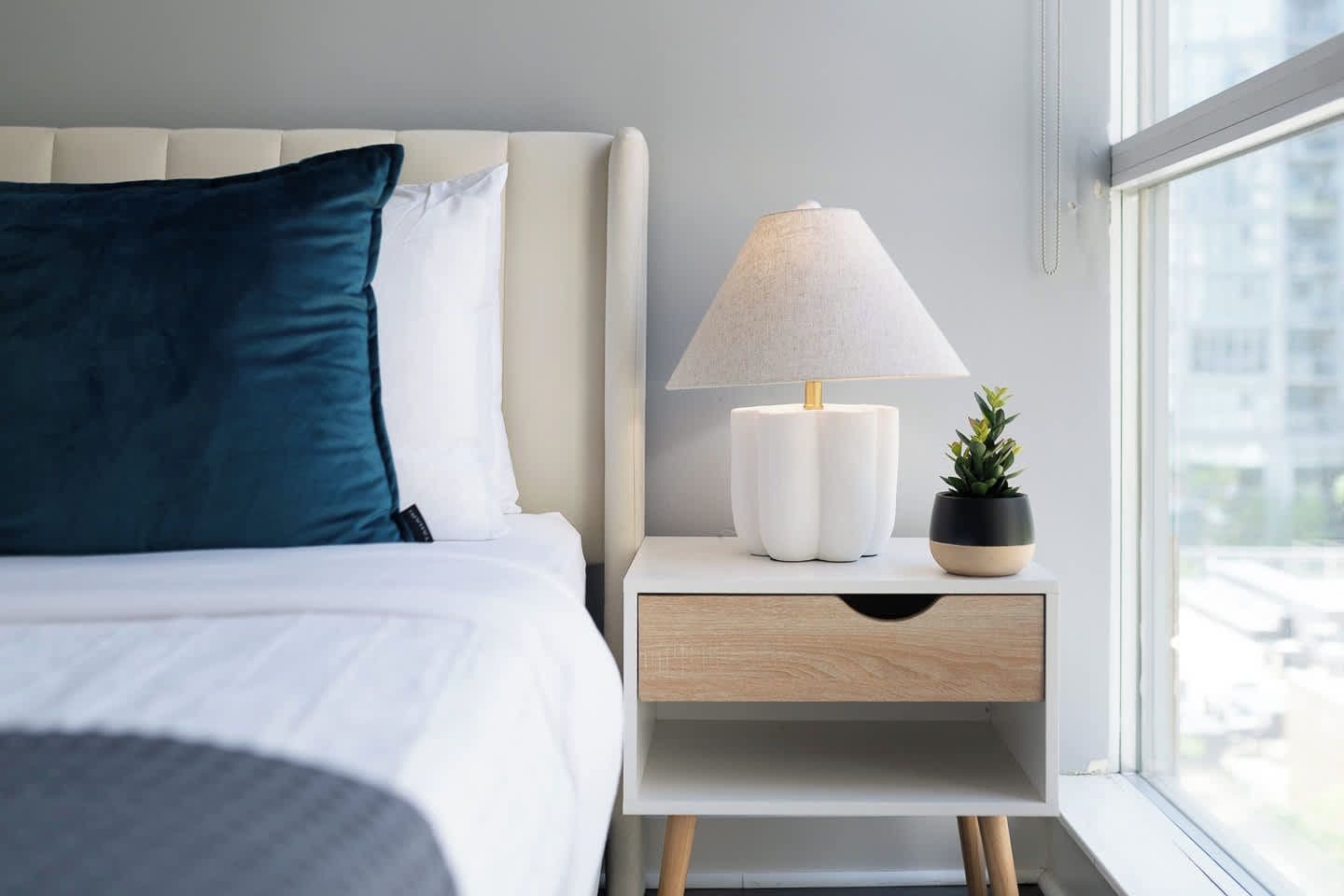 A neatly made bed with a white and blue pillow, a bedside table with a lamp and a small plant, and a window with a view of buildings.