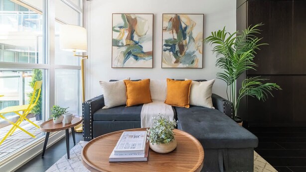 Una sala moderna con un sofá gris oscuro, dos cuadros abstractos, un sillón amarillo, una lámpara de pie y una planta en maceta.