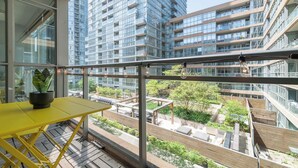 Un balcón con una mesa amarilla y una planta en maceta, con vistas a un moderno conjunto de edificios con varios pisos y balcones.
