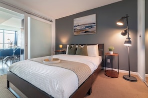 A modern bedroom with a large bed, bedside lamps, a small table with a plant, and a painting on the wall. - Spacious 3BR condo with patio & free parking (Toronto)