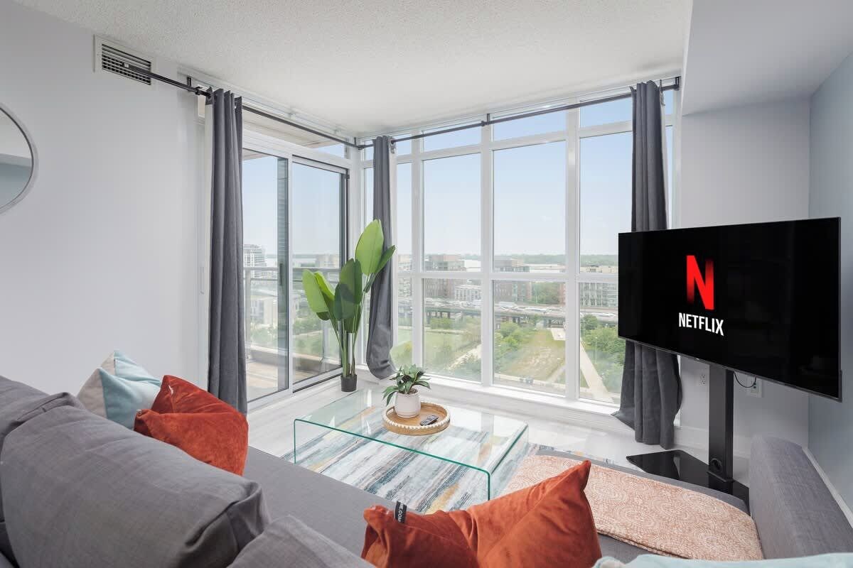 A modern living room with a large window, a flat-screen TV, and a glass coffee table.