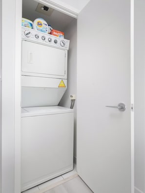 A compact, white laundry room with a washing machine and dryer stacked vertically. - Stylish 2BR Downtown Skyline Views & Free Parking | Parking Available (Toronto)