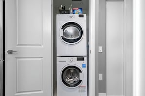 A white door with a handle, a white wall, and a white washing machine with a control panel and a detergent box on top. - Sunlit Studio w Balcony Views Prime Location! | Parking Available (Toronto)