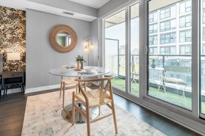 A modern dining area with a round table, two wooden chairs, a wall-mounted mirror, and a view of a building through large windows. - Sunlit Studio w Balcony Views Prime Location! | Parking Available (Toronto)