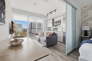 A modern apartment with a dining area, a sofa, a kitchen, and a large window offering a city view.