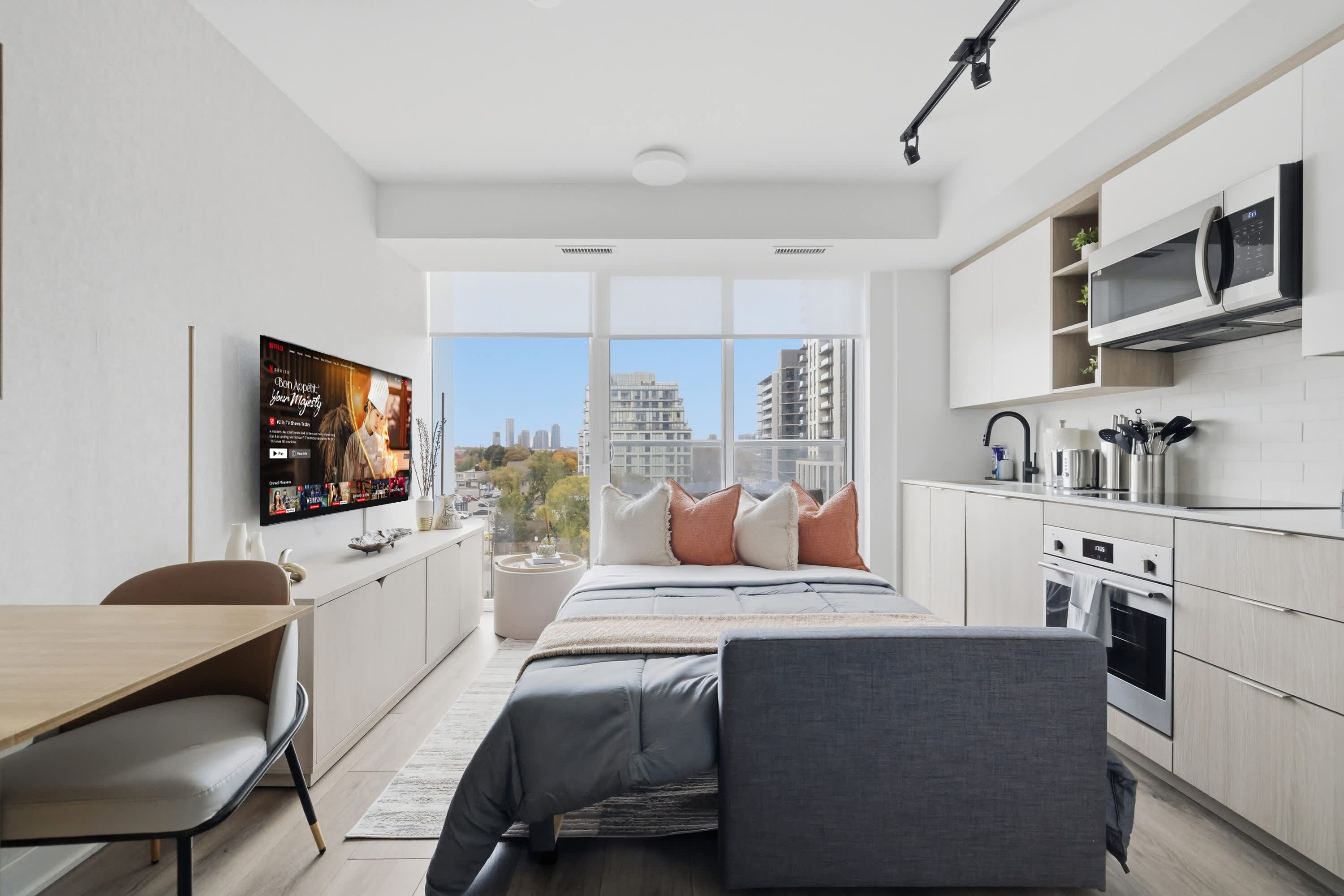 A modern hotel room with a bed, a desk, a TV, a kitchen area, and a large window offering a city view.