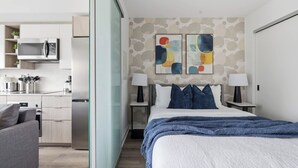 Quarto de hotel moderno com cama, mesas de cabeceira, cozinha e arte abstrata na parede.