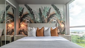 Habitación de hotel con una cama grande, cabecera, un mural de follaje tropical y lámparas de noche, con vista a la ciudad.