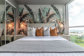 Une chambre d’hôtel avec un grand lit, une tête de lit, une fresque représentant une végétation tropicale, des lampes de chevet et une vue sur la ville.