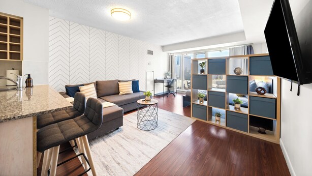 A modern living room with a herringbone-patterned wall, a herringbone-patterned sofa, a kitchen area with a granite countertop, and a large TV mounted on the wall. - Modern Downtown Studio Steps to Yonge-Dundas (Toronto)