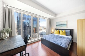A modern hotel room with a large window, a bed with blue bedding, a desk with a chair, and a painting on the wall. - Modern Downtown Studio Steps to Yonge-Dundas | Parking Available (Toronto)