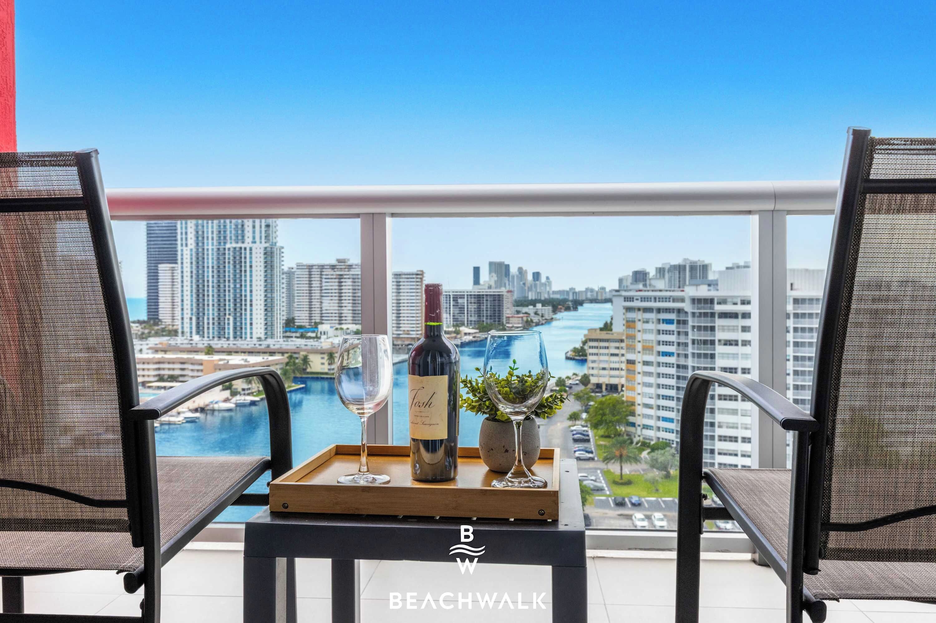 A balcony with two mesh chairs, a tray with a wine bottle and glass, and a view of a cityscape and water.