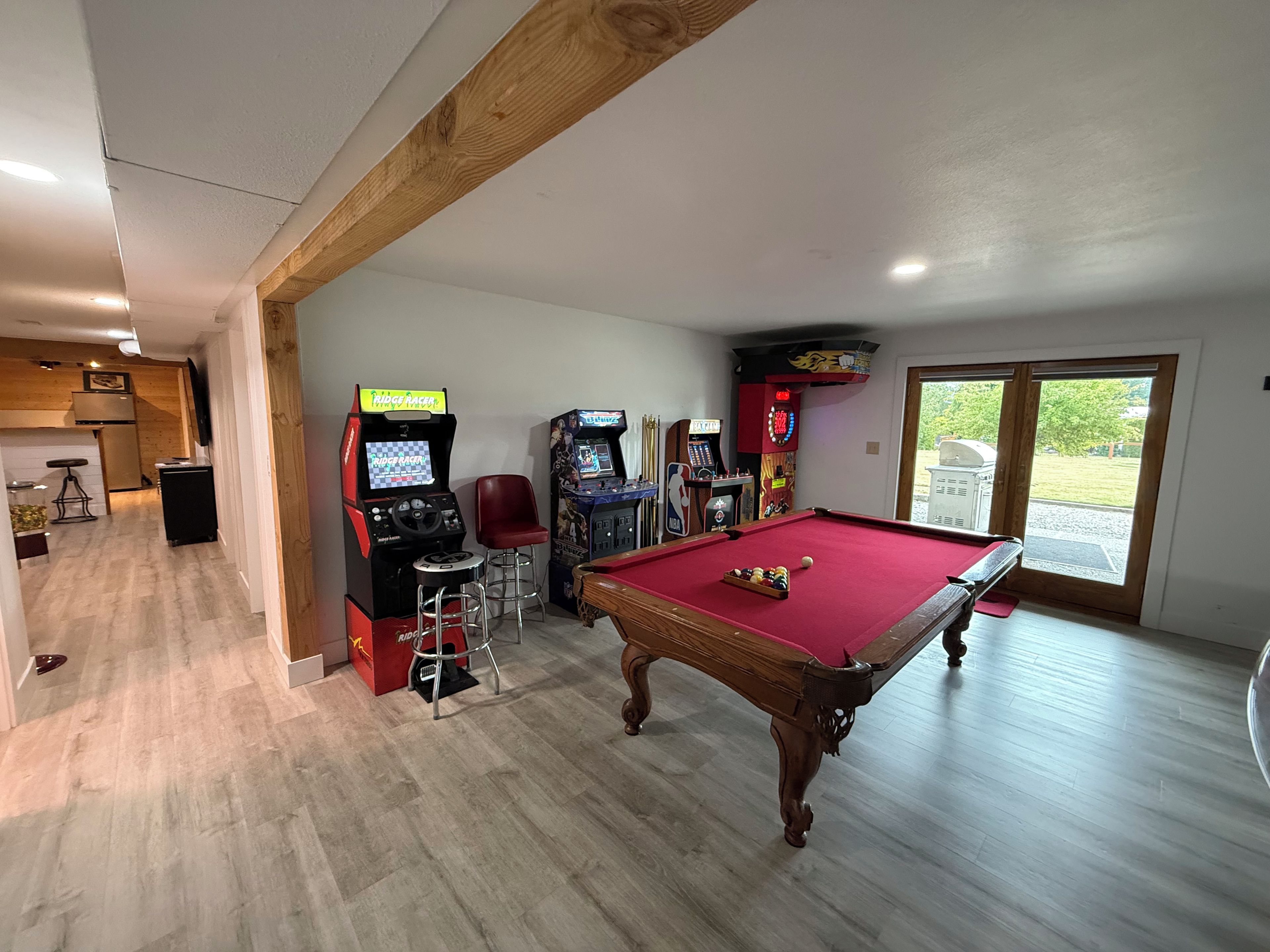 Une table de billard recouverte d’un feutre rouge, deux bornes d’arcade, un tabouret de bar rouge et une poutre en bois au plafond.