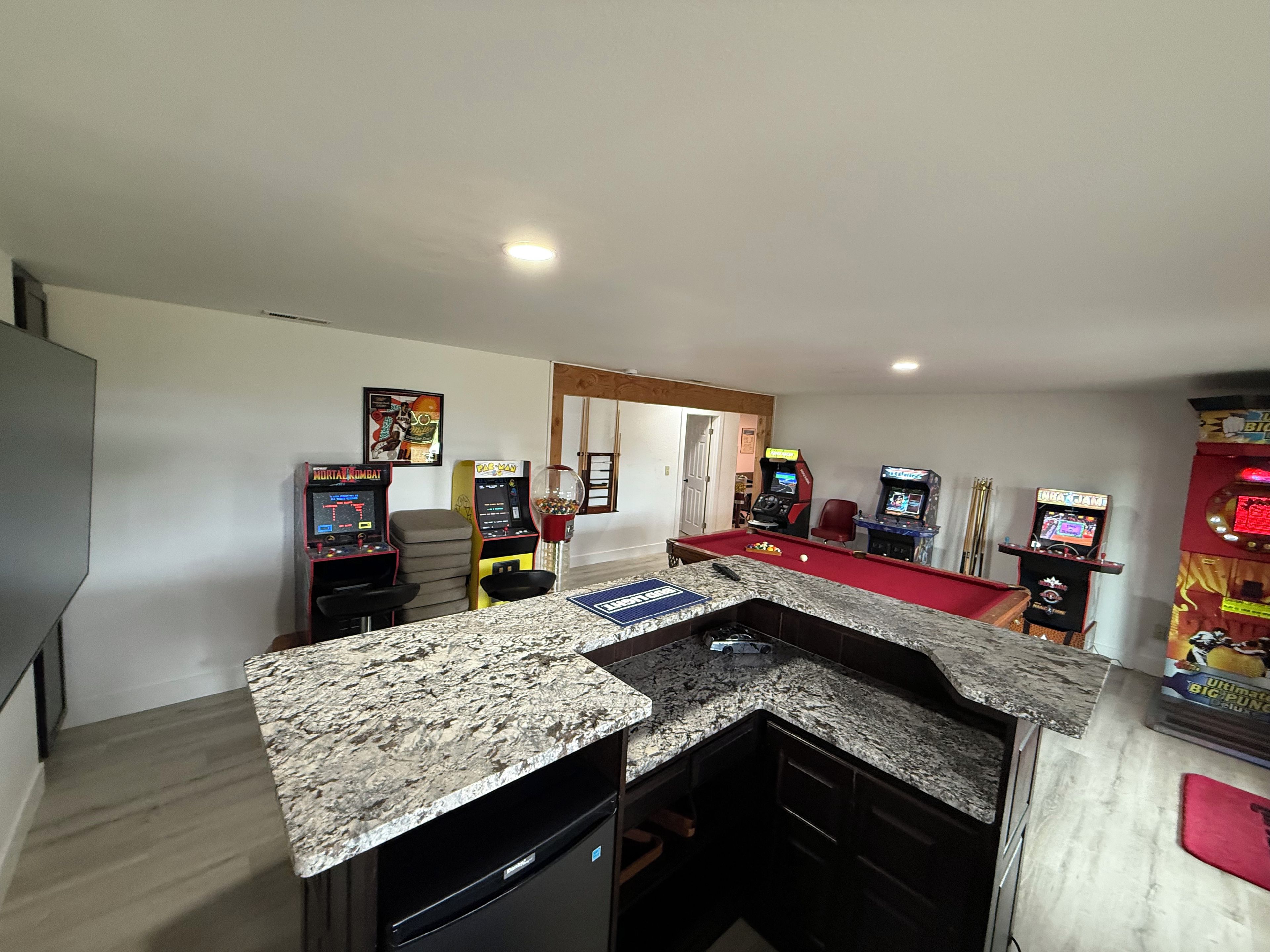 A room with a pool table, arcade games, and a kitchen area with granite countertops.