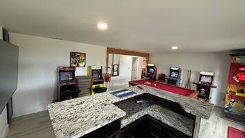 A room with a pool table, arcade games, and a kitchen area with granite countertops.