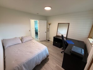A bedroom with a bed, a desk, a mirror, and an open door leading to another room.