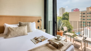 Habitación de hotel con una cama grande, una mesita con una taza y un plato, una revista, y vistas de la ciudad desde un balcón con una silla.
