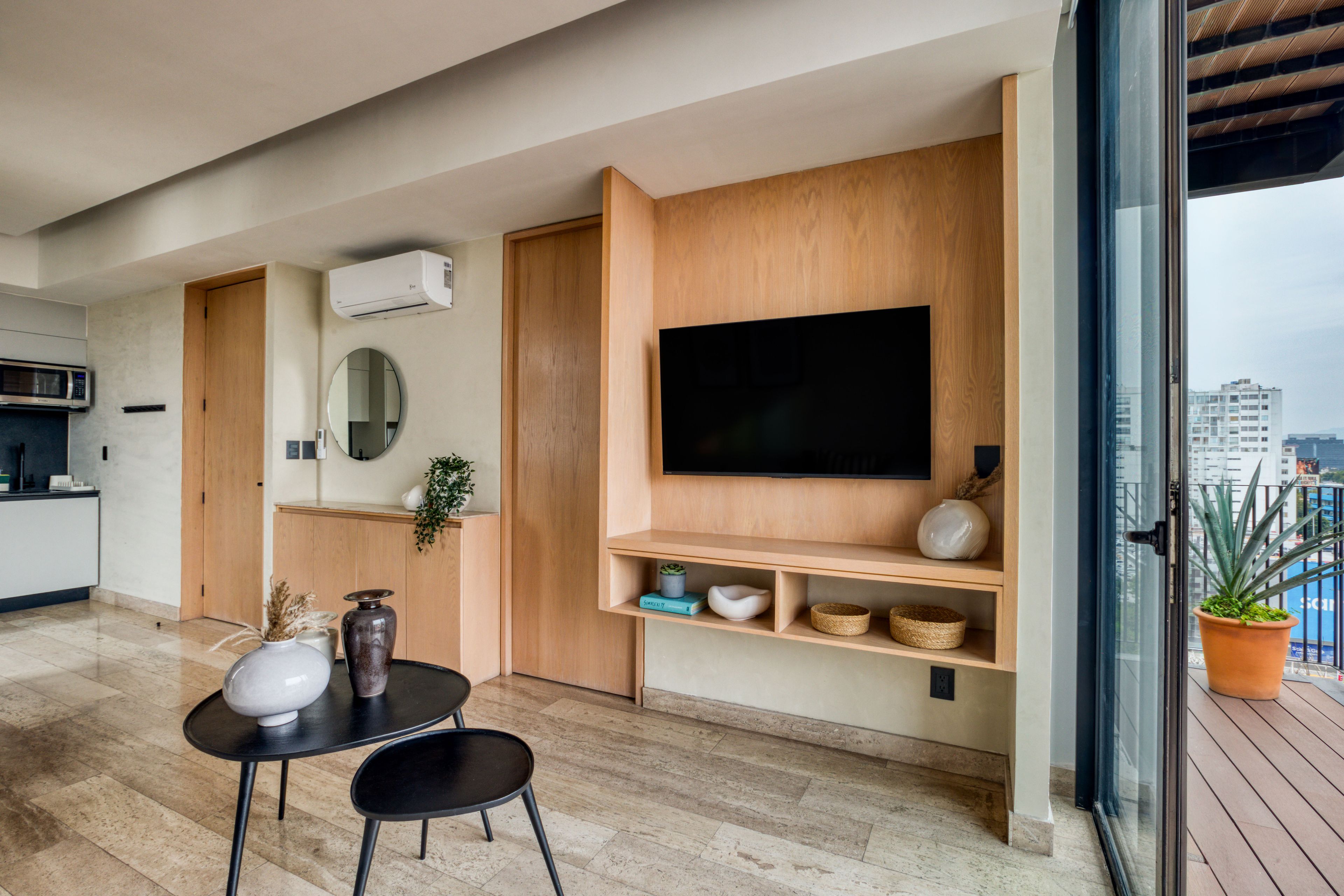 Una sala de estar moderna con un televisor de pantalla plana montado en una pared de madera, un comedor con mesa y sillas, y una planta en maceta en el balcón.