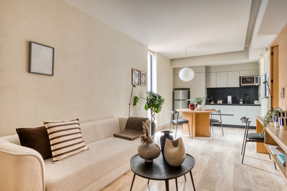 Una sala de estar moderna con un sofá, una mesa de centro con jarrones decorativos y vista al área de la cocina.