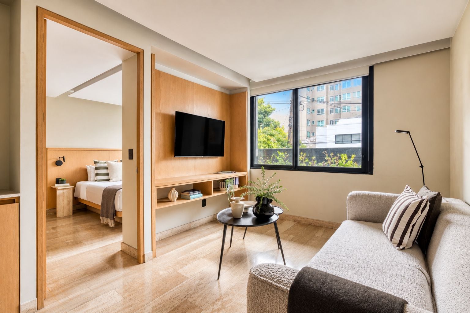 A modern living room with a sofa, a TV mounted on a wooden wall, a small table with decorative items, and a window with a view of buildings.
