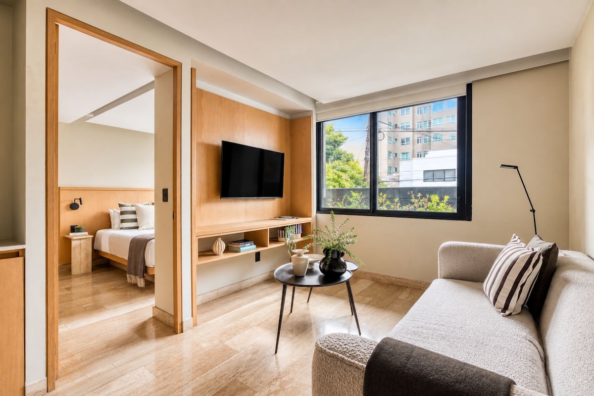 Uma sala de estar moderna com sofá, TV fixada em uma parede de madeira, uma pequena mesa com objetos decorativos e uma janela com vista para edifícios.