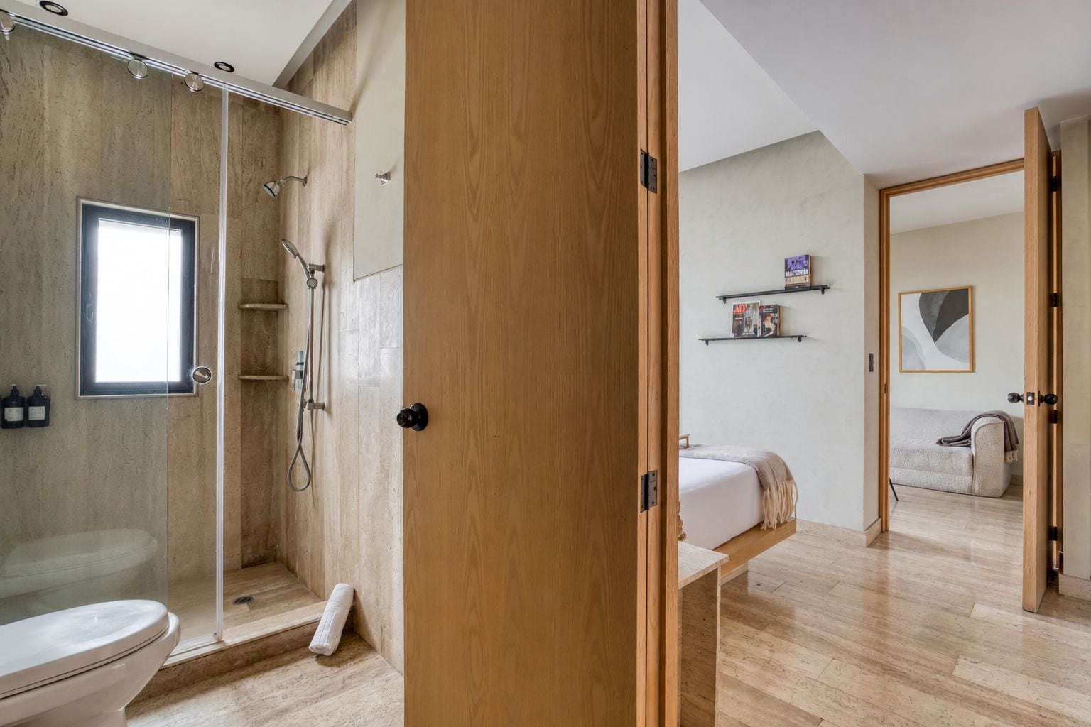 A modern bathroom with a glass shower enclosure, a toilet, and a wooden door leading to a bedroom with a bed and a nightstand.