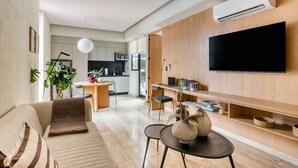 Una sala moderna con sofá, mesa de centro y televisor. Se observa un área de cocina al fondo, con microondas y mesa de comedor.