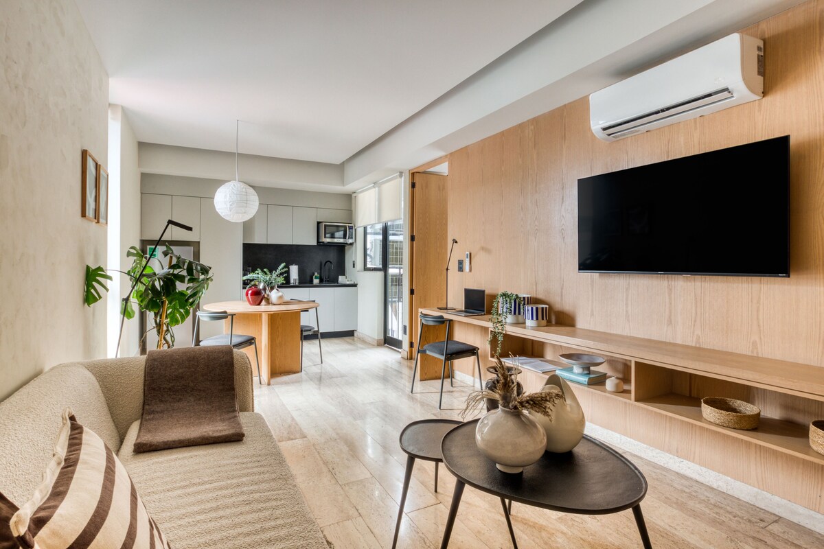 Una sala moderna con sofá, mesa de centro y televisor. Se observa un área de cocina al fondo, con microondas y mesa de comedor.