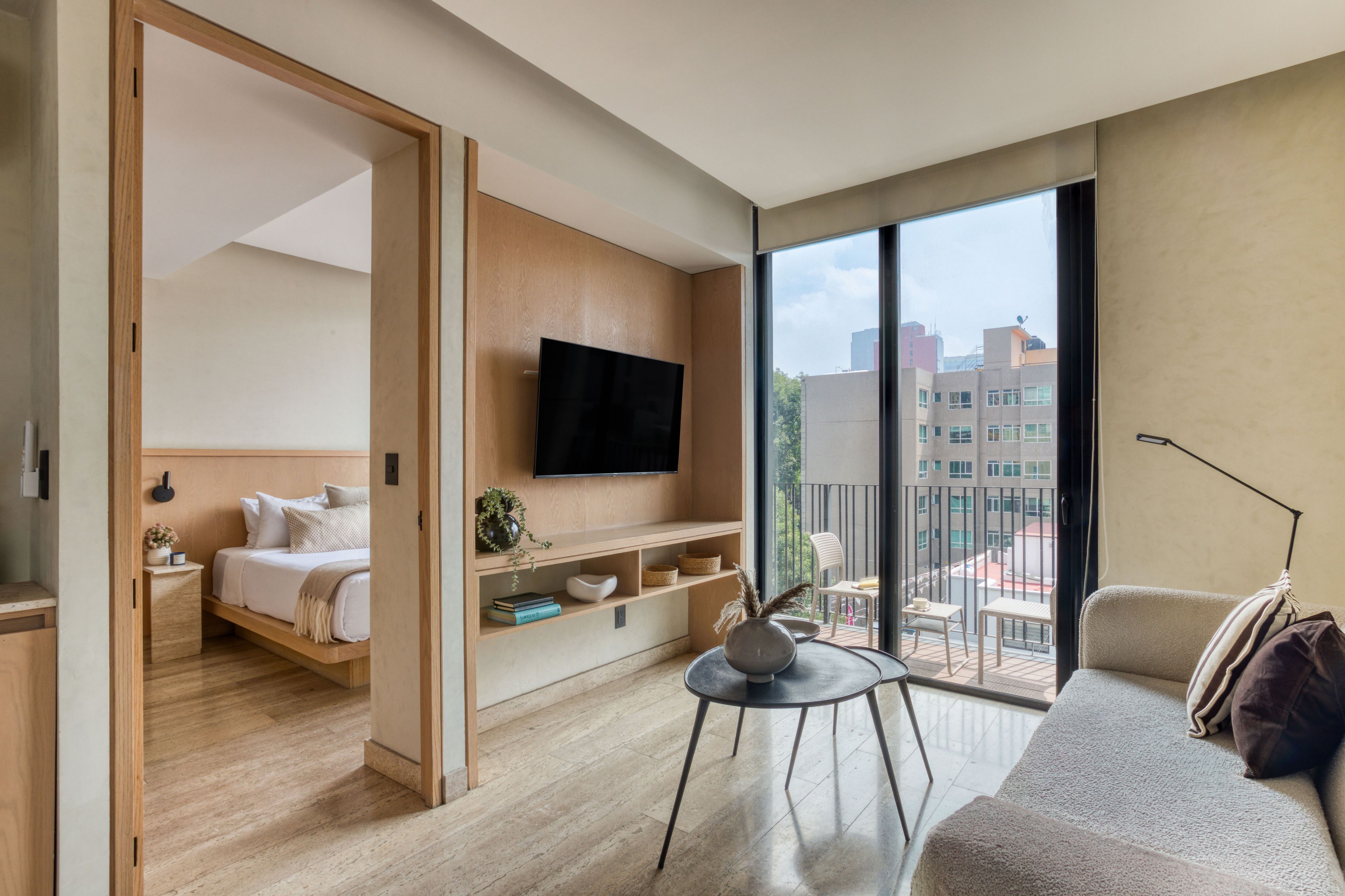 Una habitación de hotel moderna con una cama, un televisor montado en la pared, un balcón con vista a la ciudad y una zona de estar con mesa y sillas.